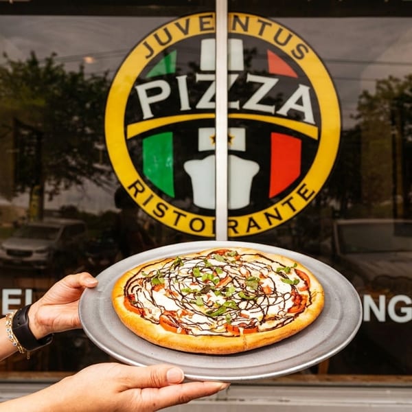 Hands holding a pizza with tomatoes, cheese, and balsamic glaze in front of a Juventus Pizza sign.