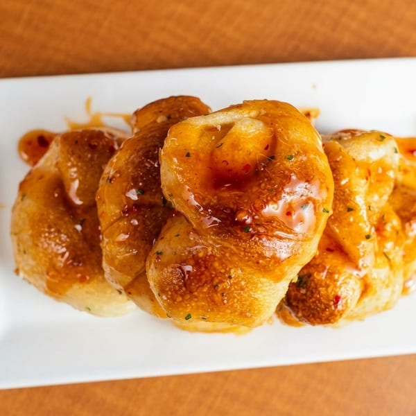 Four golden-brown garlic knots drizzled with sweet chili sauce on a white rectangular plate.